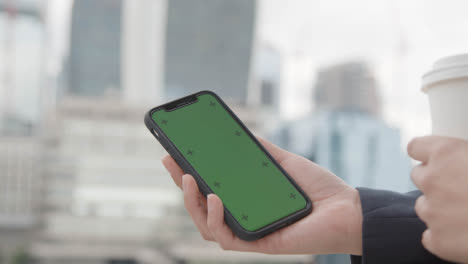 Close-Up-Of-Businessman-Holding-Takeaway-Cup-Looking-At-Green-Screen-Mobile-Phone-Outdoors