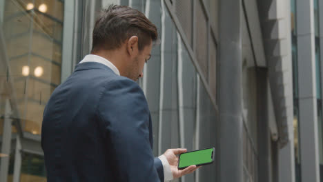 Businessman-Outside-City-Of-London-Offices-Holding-Green-Screen-Mobile-Phone-Horizontally-1