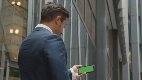 Hombre-De-Negocios-Fuera-De-Las-Oficinas-De-La-Ciudad-De-Londres-Sosteniendo-Un-Teléfono-Móvil-Con-Pantalla-Verde-Horizontalmente-1