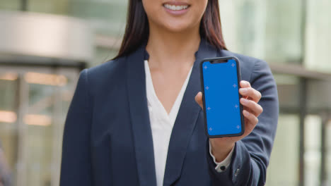 Close-Up-Of-Businesswoman-Outside-City-Of-London-Offices-Holding-Blue-Screen-Mobile-Phone