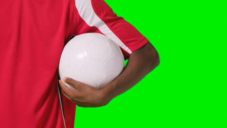 Nahaufnahme-Studioaufnahme-Eines-Männlichen-Fußballers,-Der-Club-Kit-Mit-Ball-Unter-Dem-Arm-Vor-Grünem-Bildschirm-Trägt