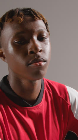 Vertical-Video-Studio-Portrait-Of-Young-Male-Footballer-Wearing-Club-Kit-With-Ball-Under-Arm-