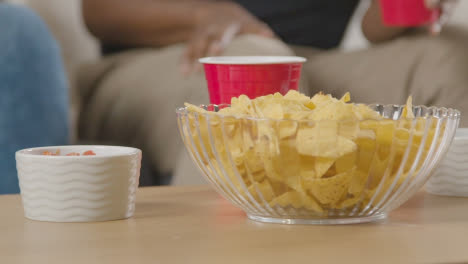 Cerca-De-Una-Pareja-Sentada-En-Un-Sofá-En-Casa-Juntos-Comiendo-Papas-Fritas-Y-Salsas