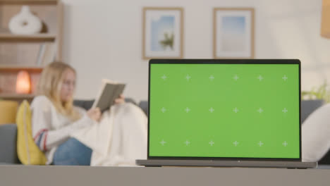 Woman-Relaxing-On-Sofa-Reading-Book-At-Home-With-Green-Screen-Laptop-In-Foreground-