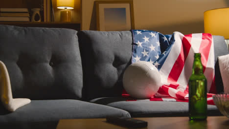 Sofa-In-Der-Lounge-Mit-Amerikanischer-Flagge-Und-Ball,-Während-Sich-Die-Fans-Darauf-Vorbereiten,-Fußballspiele-Im-Fernsehen-Zu-Sehen