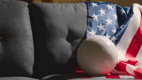 Sofa-In-Der-Lounge-Mit-Amerikanischer-Flagge-Und-Ball,-Während-Sich-Die-Fans-Darauf-Vorbereiten,-Fußballspiele-Im-Fernsehen-1-Zu-Sehen