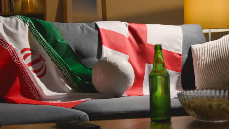 Sofa-In-Der-Lounge-Mit-Englischen-Und-Iranischen-Flaggen-Und-Ball,-Während-Fans-Fußballspiele-Im-Fernsehen-Sehen