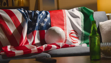 Sofa-In-Der-Lounge-Mit-Iranischen-Und-Amerikanischen-Flaggen-Und-Ball,-Während-Fans-Fußballspiele-Im-Fernsehen-1-Sehen