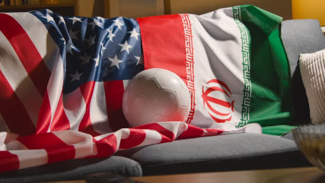 Sofa-In-Der-Lounge-Mit-Iranischen-Und-Amerikanischen-Flaggen-Und-Ball,-Während-Fans-Fußballspiele-Im-Fernsehen-Sehen-2