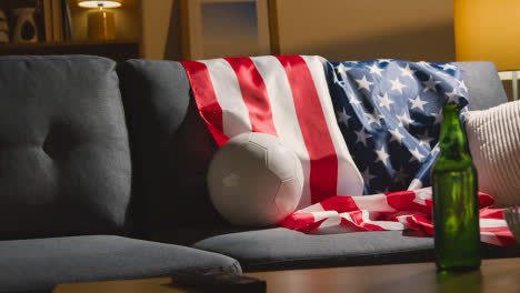 Sofa-In-Der-Lounge-Mit-Amerikanischer-Flagge-Und-Ball,-Während-Sich-Die-Fans-Darauf-Vorbereiten,-Fußballspiele-Im-Fernsehen-Zu-Sehen-2