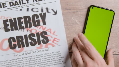 Zeitung-Mit-Schlagzeile-Zur-Energiekrise-Neben-Der-Hand,-Die-Das-Handy-Mit-Grünem-Bildschirm-In-Die-Hand-Nimmt