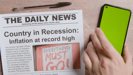 Zeitung-Mit-Schlagzeile-über-Rezession-Neben-Der-Hand,-Die-Das-Handy-Mit-Grünem-Bildschirm-Aufhebt