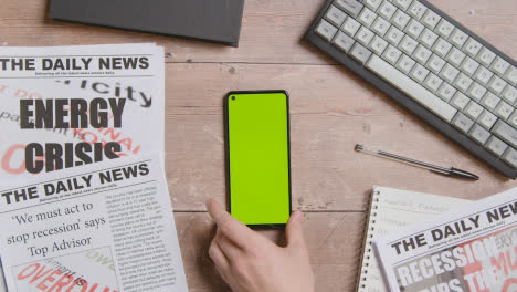 Overhead-Shot-Of-Table-With-Hand-Picking-Up-Green-Screen-Mobile-Phone-With-Newspaper-Stories-On-Recession-And-Energy-Crisis