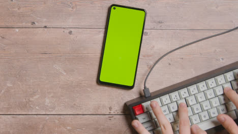 Fotografía-Cenital-De-La-Mesa-Y-Las-Manos-Escribiendo-En-El-Teclado-De-La-Computadora-Con-El-Teléfono-Móvil-De-Pantalla-Verde-1