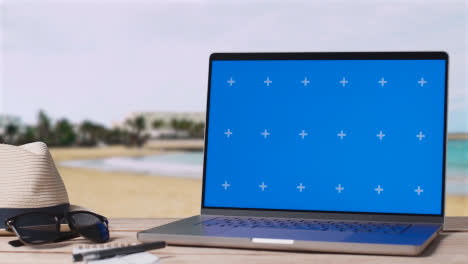 Blue-Screen-Laptop-On-Table-With-Holiday-Beach-And-Hotel-In-Background