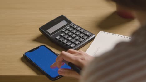 Woman-Using-Blue-Screen-Mobile-Phone-And-Calculator-To-Check-Household-Budget-In-Cost-Of-Living-Crisis