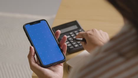 Woman-Using-Blue-Screen-Mobile-Phone-And-Calculator-To-Check-Household-Budget-In-Cost-Of-Living-Crisis-1