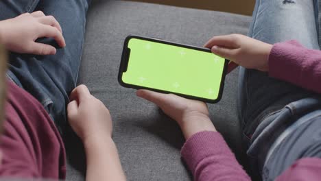 Primer-Plano-De-Dos-Niños-Sentados-En-Un-Sofá-En-Casa-Mirando-Un-Teléfono-Móvil-De-Pantalla-Verde