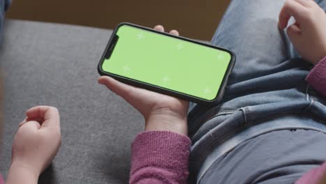 Primer-Plano-De-Dos-Niños-Sentados-En-Un-Sofá-En-Casa-Mirando-El-Teléfono-Móvil-De-Pantalla-Verde-3