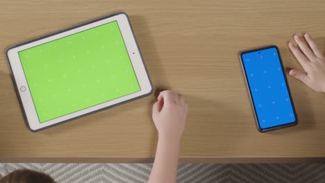 Überkopfaufnahme-Von-Kindern,-Die-Zu-Hause-Ein-Digitales-Tablet-Mit-Grünem-Bildschirm-Und-Ein-Handy-Mit-Blauem-Bildschirm-Auf-Dem-Tisch-Verwenden