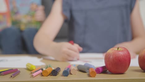 Nahaufnahme-Von-Kindern-Zu-Hause,-Die-Ein-Bild-Von-Essen-Am-Tisch-Ausmalen-Und-Ein-Buch-Mit-Frischem-Apfel-Im-Vordergrund-Lesen