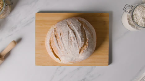 Overhead-Shot-Of-Freshly-Baked-Loaf-Of-Bread-Being-Picked-Up-From-Wooden-Board