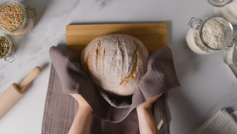 Overhead-Shot-Of-Freshly-Baked-Loaf-Of-Bread-Being-Put-Down-Onto-Wooden-Board