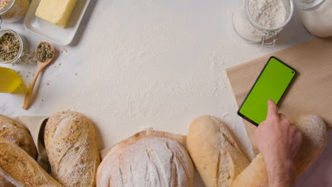 Fotografía-Cenital-De-Hogazas-De-Pan-Recién-Horneadas-En-Una-Superficie-De-Trabajo-De-Mármol-Con-Un-Teléfono-Móvil-De-Pantalla-Verde