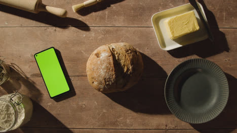 Fotografía-Cenital-De-Una-Hogaza-De-Pan-Recién-Horneada-Sobre-Una-Mesa-De-Madera-Con-Un-Teléfono-Móvil-De-Pantalla-Verde-1