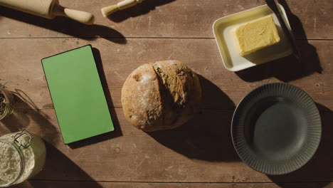 Fotografía-Cenital-De-Una-Hogaza-De-Pan-Recién-Horneada-Sobre-Una-Mesa-De-Madera-Con-Una-Tableta-Digital-De-Pantalla-Verde