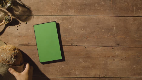 Fotografía-Cenital-De-Una-Persona-Recogiendo-Una-Hogaza-De-Pan-De-Una-Mesa-De-Madera-Con-Una-Tableta-Digital-De-Pantalla-Verde