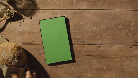 Fotografía-Cenital-De-Una-Persona-Que-Deja-Una-Hogaza-De-Pan-De-Una-Mesa-De-Madera-Con-Una-Tableta-Digital-De-Pantalla-Verde