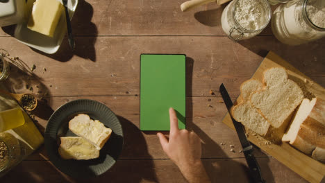 Fotografía-Cenital-De-Una-Persona-Comiendo-Pan-Recién-Horneado-Con-Mantequilla-En-Una-Mesa-De-Madera-Con-Una-Tableta-Digital-De-Pantalla-Verde