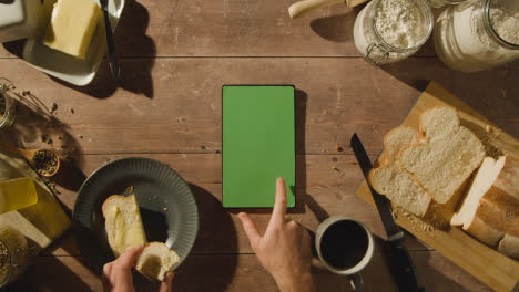 Fotografía-Cenital-De-Una-Persona-Comiendo-Pan-Recién-Horneado-Con-Mantequilla-En-Una-Mesa-De-Madera-Con-Una-Tableta-Digital-De-Pantalla-Verde-1