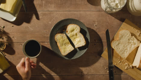 Fotografía-Cenital-De-Una-Persona-Comiendo-Pan-Recién-Horneado-Con-Mantequilla-En-Una-Mesa-De-Madera-Con-Bebida-Caliente