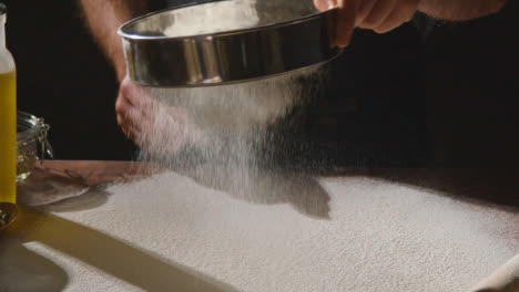 Low-Key-Shot-Of-Person-Sieving-Flour-Onto-Work-Surface-For-Baking-With-Ingredients