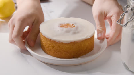 Person-Picking-Up-Homemade-Lemon-Drizzle-Cake-On-Plate-On-Kitchen-Work-Surface-Behind-Ingredients-1