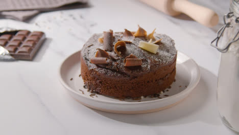 Person-Putting-Homemade-Chocolate-Cake-On-Plate-Onto-Kitchen-Work-Surface-With-Ingredients-1