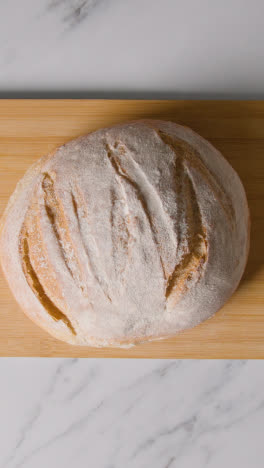 Vertical-Video-Overhead-Shot-Of-Freshly-Baked-Loaf-Of-Bread-Being-Picked-Up-From-Wooden-Board