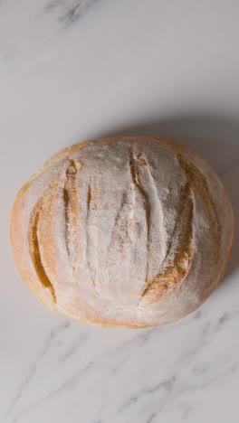 Vertical-Video-Overhead-Shot-Of-Freshly-Baked-Loaf-Of-Bread-Being-Put-Down-Onto-Marble-Work-Surface