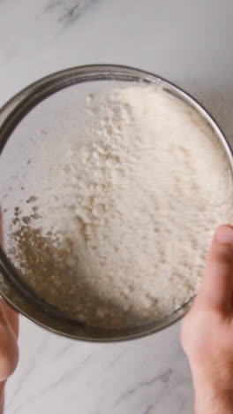 Vertical-Video-Overhead-Shot-Of-Flour-Being-Sieved-Onto-Marble-Work-Surface-For-Baking