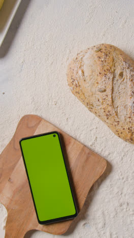 Vertical-Video-Overhead-Shot-Of-Person-Putting-Loaf-Of-Bread-Onto-Floured-Kitchen-Surface-With-Green-Screen-Mobile-Phone