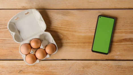 Overhead-Shot-Of-Six-Eggs-In-Cardboard-Box-Being-Opened-On-Wooden-Table-With-Mobile-Phone
