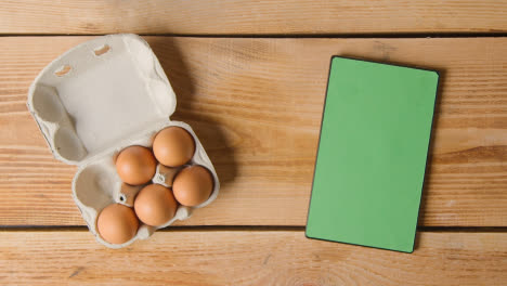 Fotografía-Cenital-De-Seis-Huevos-En-Una-Caja-De-Cartón-Que-Se-Abre-Sobre-Una-Mesa-De-Madera-Con-Una-Tableta-Digital