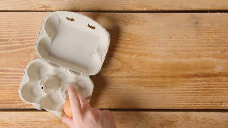 Fotografía-Cenital-De-La-Mano-Sacando-El-Huevo-De-La-Caja-De-Cartón-En-La-Mesa-De-Madera