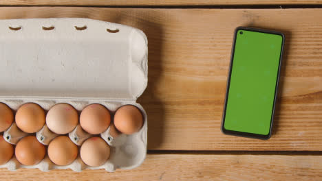Overhead-Shot-Of-Person-Choosing-Egg-From-Cardboard-Box-On-Wooden-Table-With-Mobile-Phone