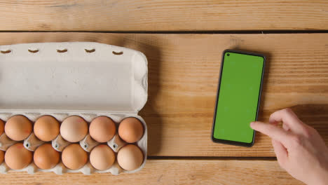 Overhead-Shot-Of-Person-Following-Recipe-On-Green-Screen-Mobile-Phone-With-Box-Of-Eggs-On-Wooden-Table