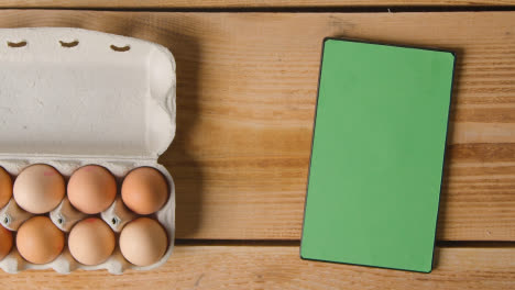 Overhead-Shot-Of-Six-Eggs-In-Open-Cardboard-Box-On-Wooden-Table-With-Digital-Tablet