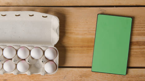 Overhead-Shot-Of-Person-Choosing-Egg-From-Cardboard-Box-On-Wooden-Table-With-Digital-Tablet