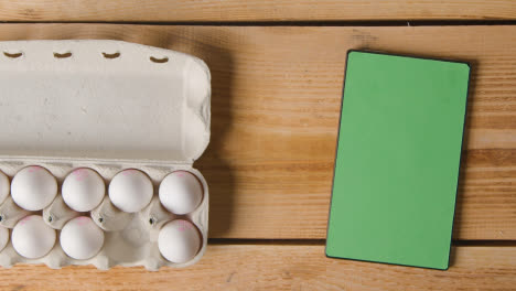 Fotografía-Cenital-De-Una-Persona-Que-Elige-Un-Huevo-De-Una-Caja-De-Cartón-Sobre-Una-Mesa-De-Madera-Con-Una-Tableta-Digital